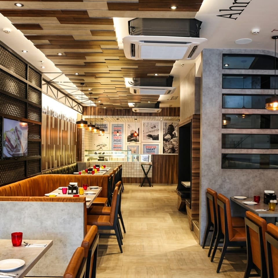 Modern restaurant interior with booths, tables, and pendant lights, featuring a wooden ceiling and city names on the wall.