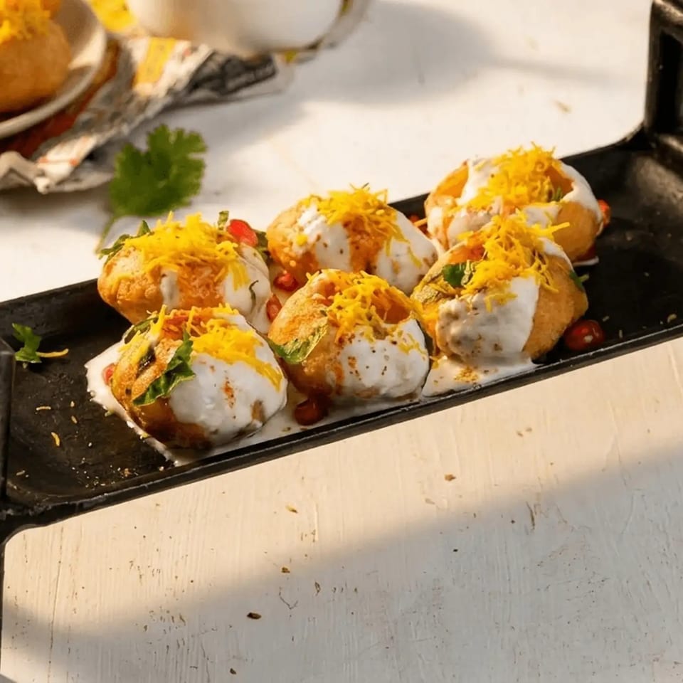 Six crispy pani puri topped with yogurt, sev, herbs, and chutney arranged on a black serving stand.