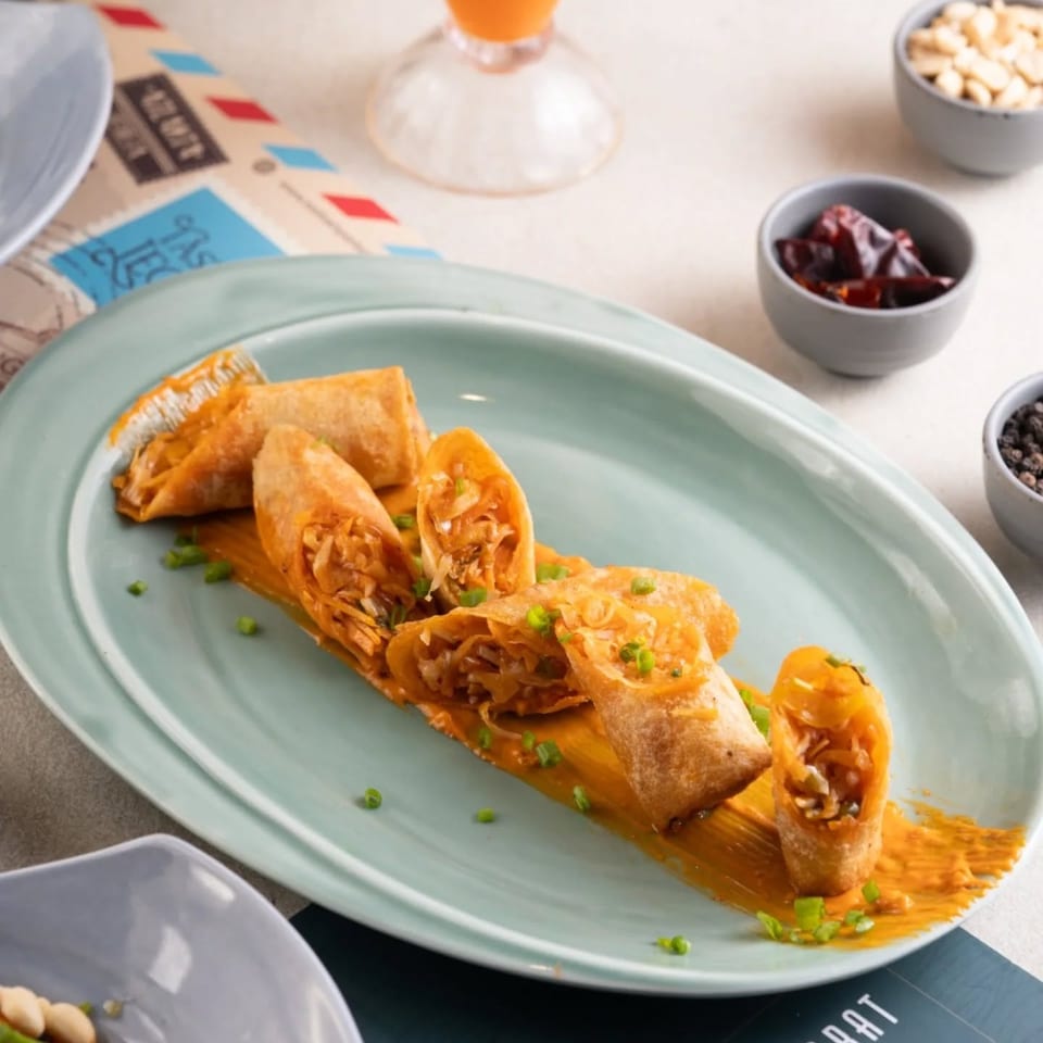 Crispy spring rolls filled with vegetables sit on orange sauce, garnished with scallions, alongside small bowls of toppings and an orange drink.