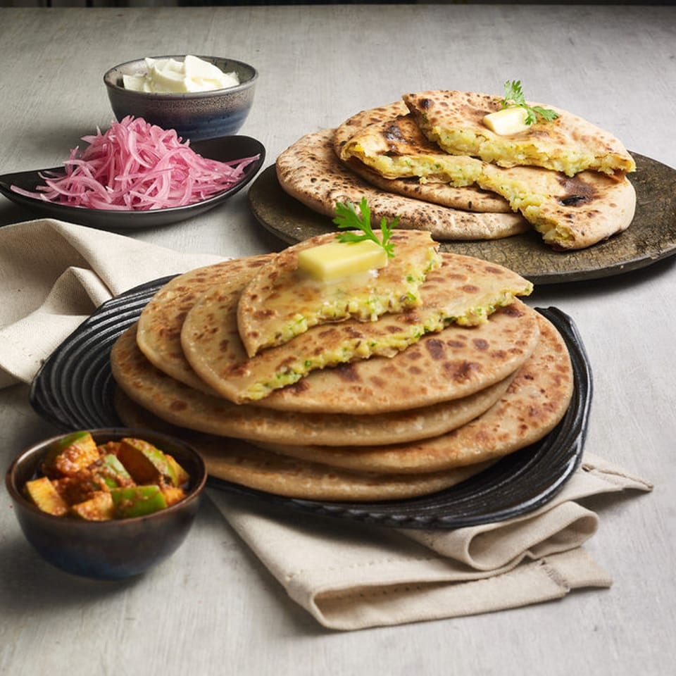 Stacked parathas with butter, pickled onions, yogurt, and mango pickle on a table.