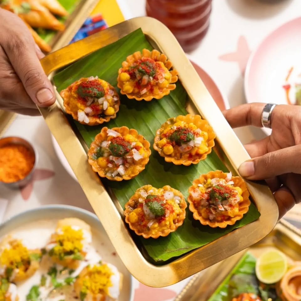 Hands present a tray of savory Indian tartlets topped with chickpeas, onions, herbs, and spices over a banana leaf.