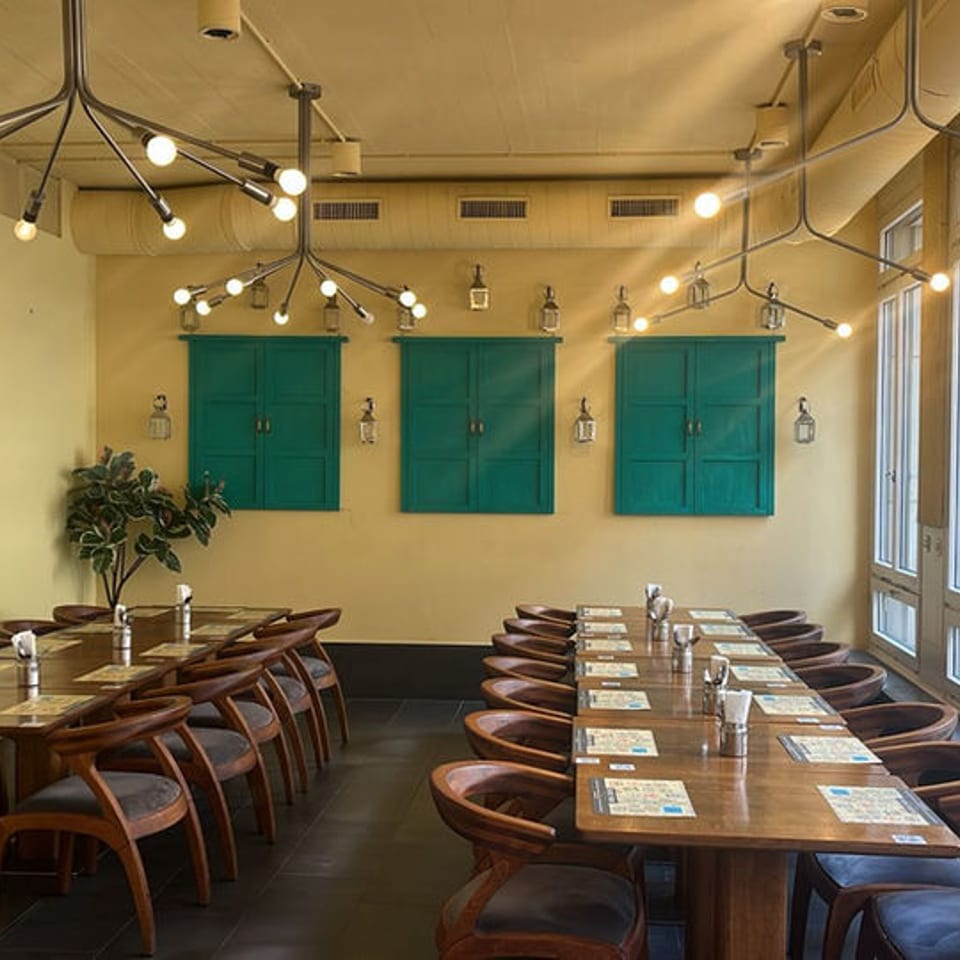 Restaurant interior with two long wooden tables, yellow walls, teal shutters, and modern lighting.